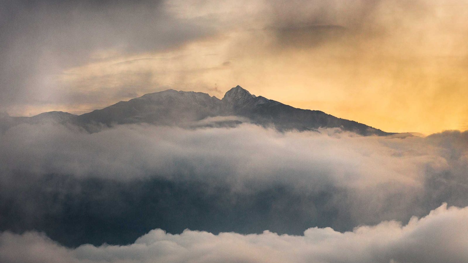 Cette photo capture la célèbre Croix du Canigó, perchée au sommet de la majestueuse montagne Canigó, dans les Pyrénées Orientales. La croix, richement décorée de rubans et de drapeaux colorés, symbolise à la fois un lieu spirituel et un point de rencontre pour les randonneurs. Les tissus flottant au vent ajoutent une touche de mouvement et de vie à la scène. Le panorama est époustouflant, avec une mer de nuages couvrant la vallée en contrebas, tandis que les crêtes montagneuses se découpent à l'horizon. La lumière douce du soleil, soit au lever soit au coucher, baigne la scène dans des teintes dorées, accentuant les reliefs rocheux qui entourent la croix. Cette image illustre parfaitement l’âme du Canigó, montagne sacrée des Catalans.