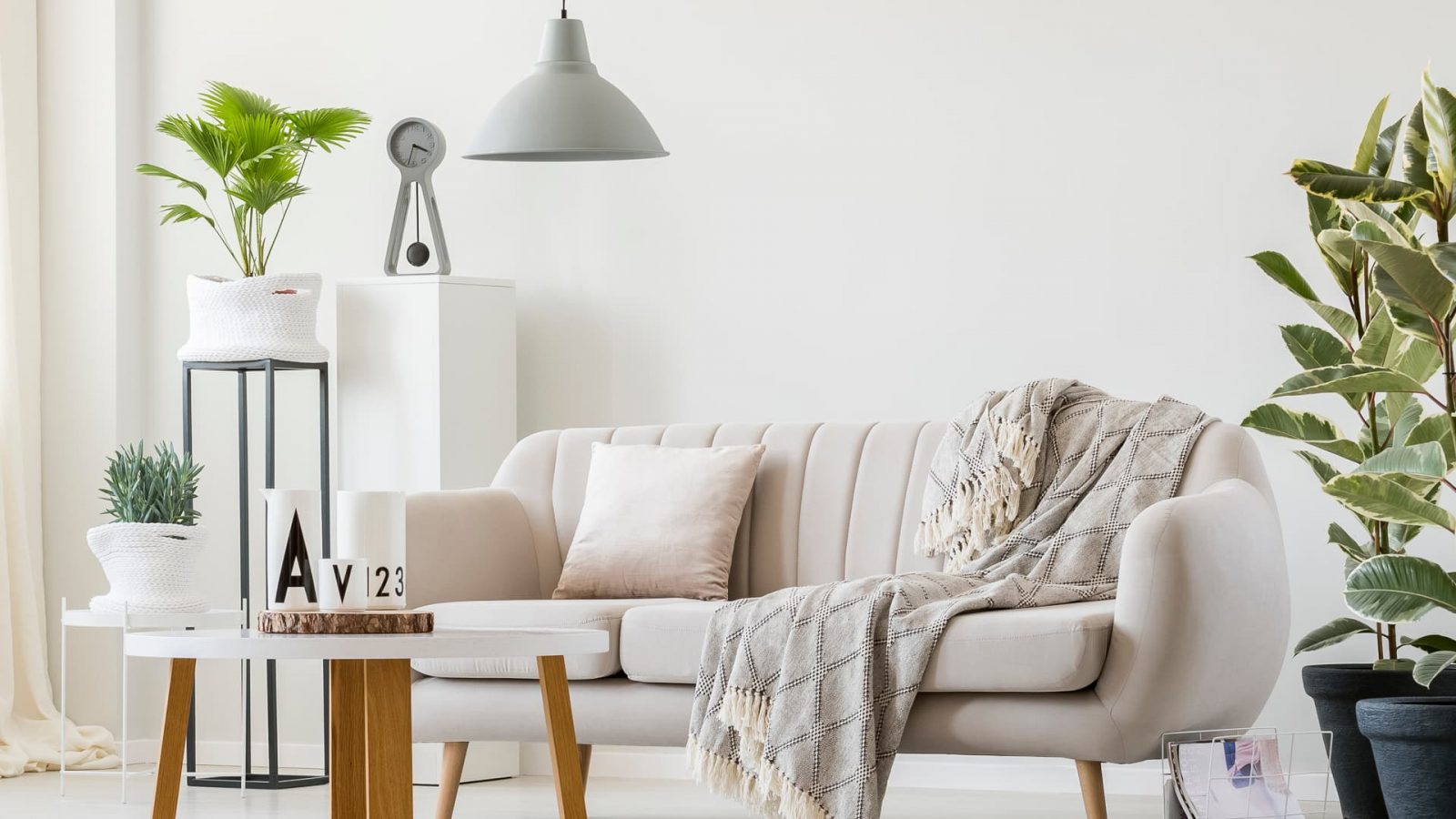 L'image montre un intérieur moderne et minimaliste avec un design épuré. Au centre, un canapé en tissu beige clair avec des lignes douces et des pieds en bois, décoré d'un coussin et d'une couverture à motifs en losanges. Devant le canapé se trouve une table basse ronde avec un plateau blanc et des pieds en bois, ornée de quelques éléments décoratifs comme des vases blancs et un petit pot de plantes. À gauche, un grand pot en tissu blanc contenant une plante verte est posé sur un support en métal noir. À droite, de grandes plantes en pot ajoutent une touche de verdure à l'ensemble de l'espace, créant une atmosphère naturelle et apaisante. Le tout est éclairé par une suspension de style industriel au-dessus de la scène, tandis que la palette de couleurs neutres avec des touches de bois et de verdure crée une ambiance harmonieuse et cosy. Cette pièce dégage une sensation de calme et de détente, parfaite pour un espace de vie confortable et moderne.